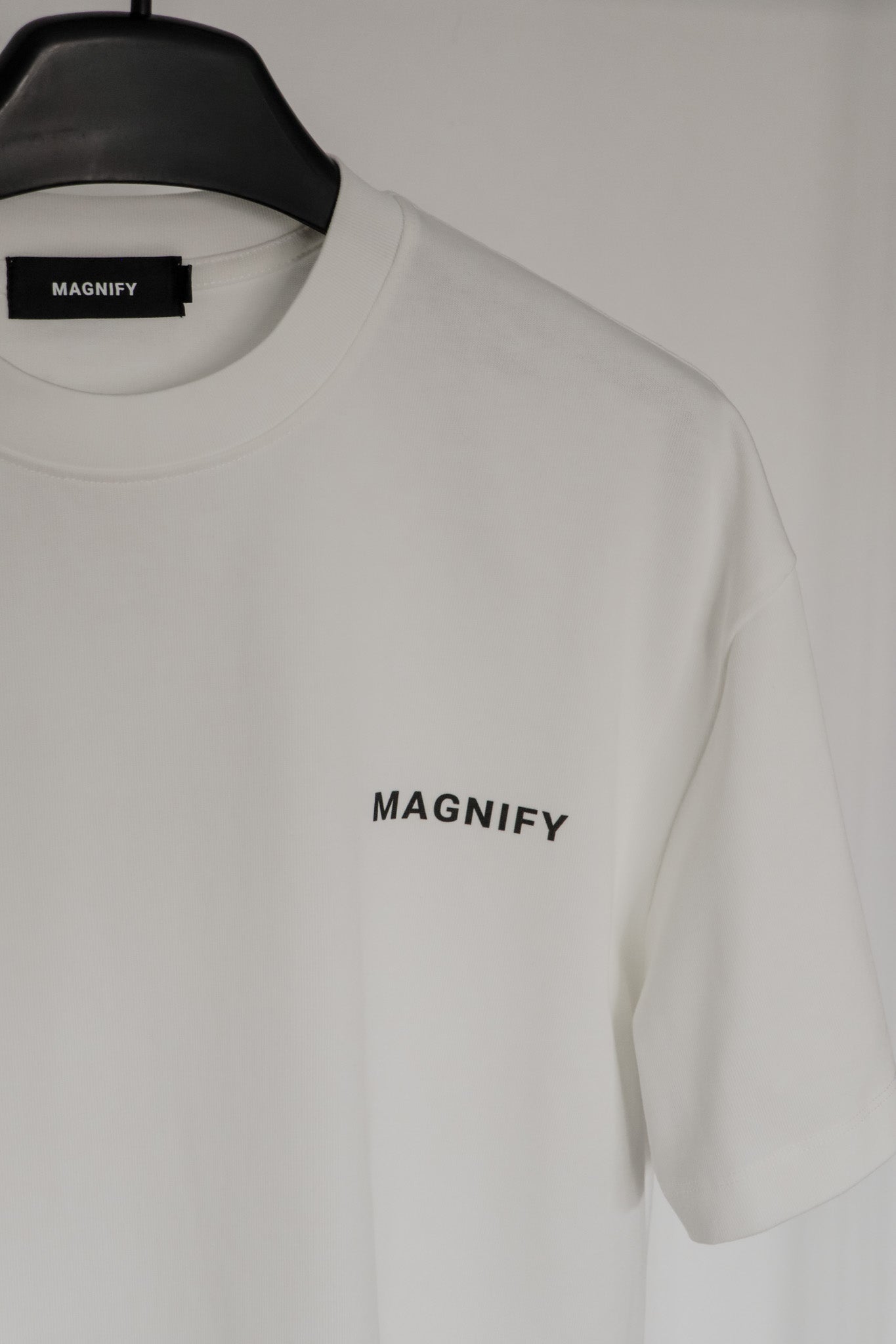 White t-shirt with 'MAGNIFY' logo on a hanger against a white background