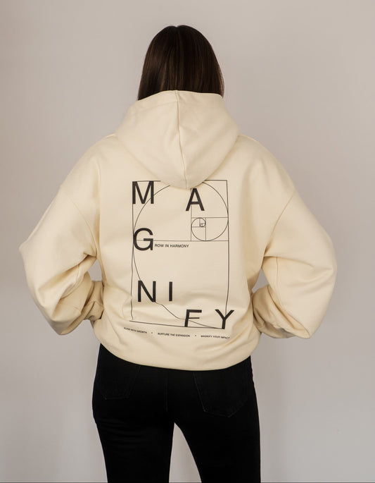 Person wearing a beige hoodie with 'MAGNIFY' design on the back against a plain background