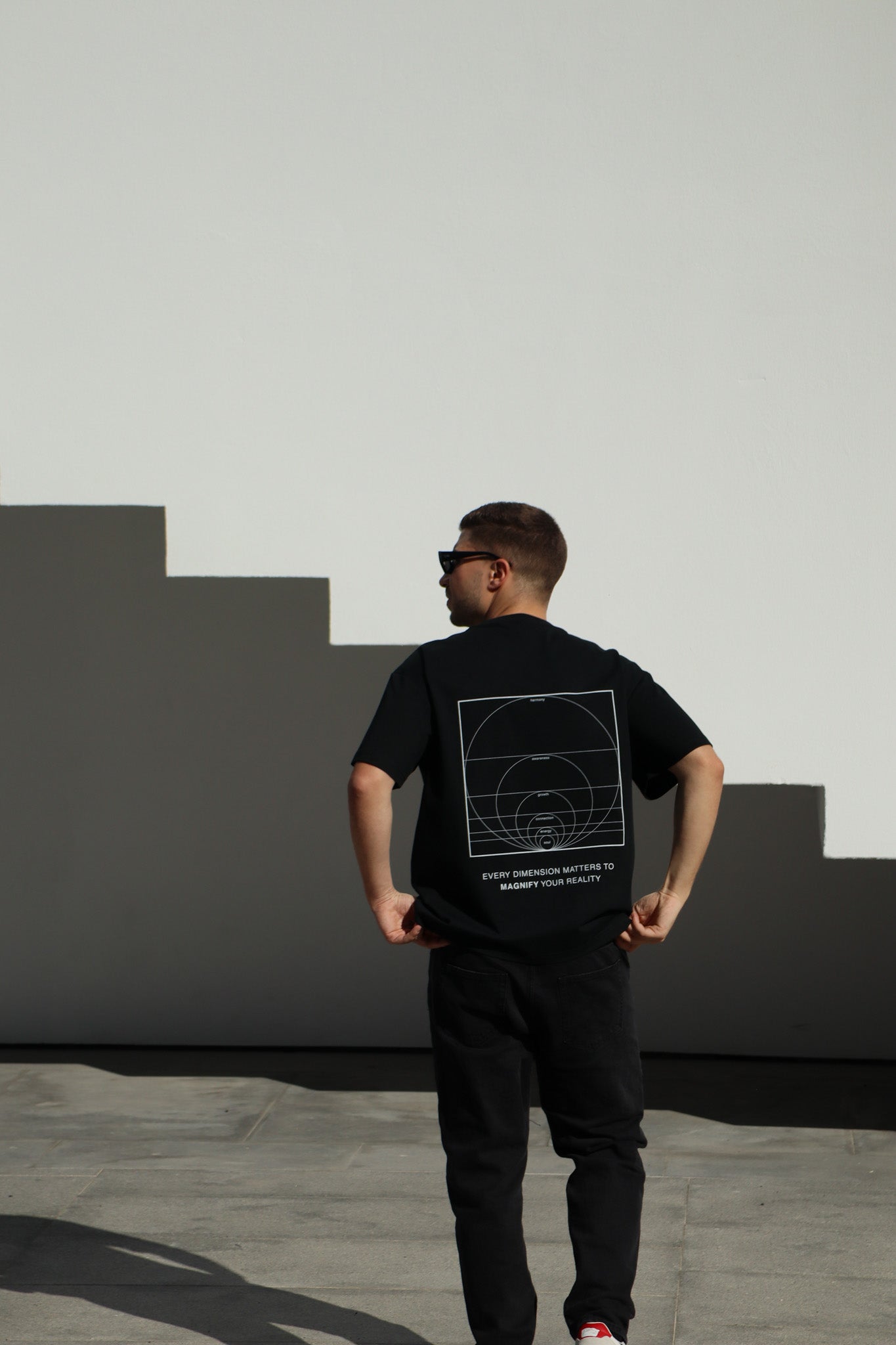 Person wearing a black t-shirt with a geometric design on the back, standing against a light-colored wall.
