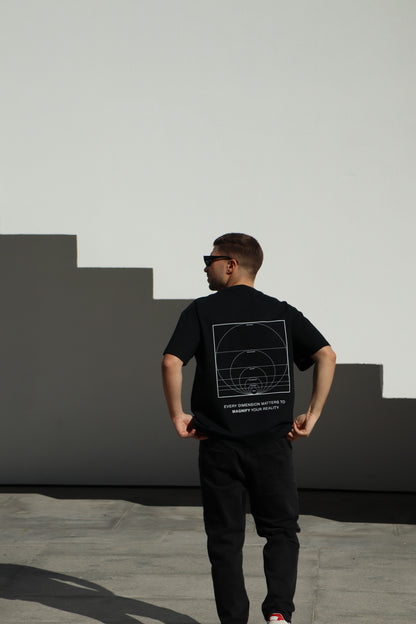 Person wearing a black t-shirt with a geometric design on the back, standing against a light-colored wall.