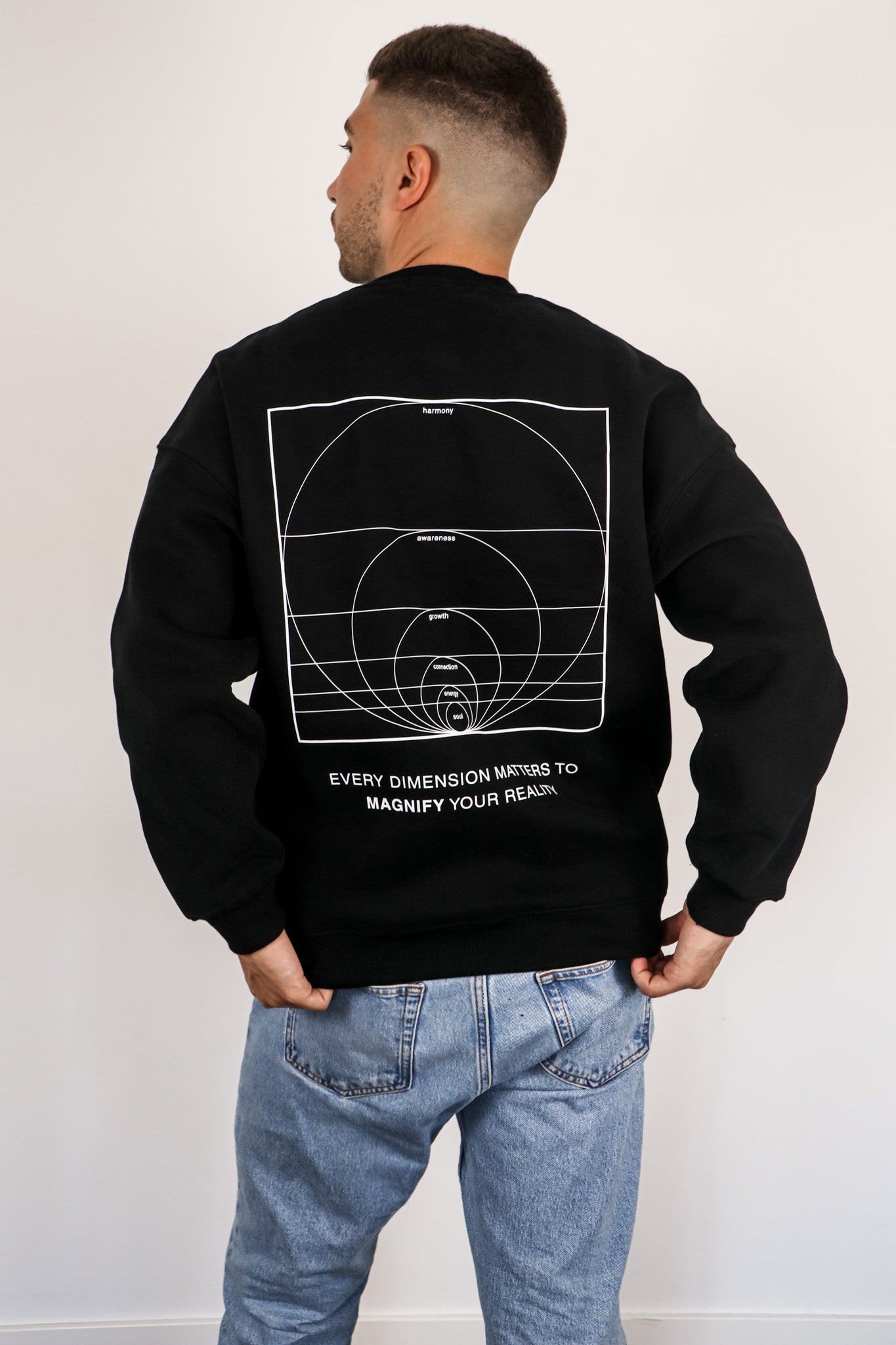 Person wearing a black sweatshirt with a geometric design on the back, standing against a white wall.