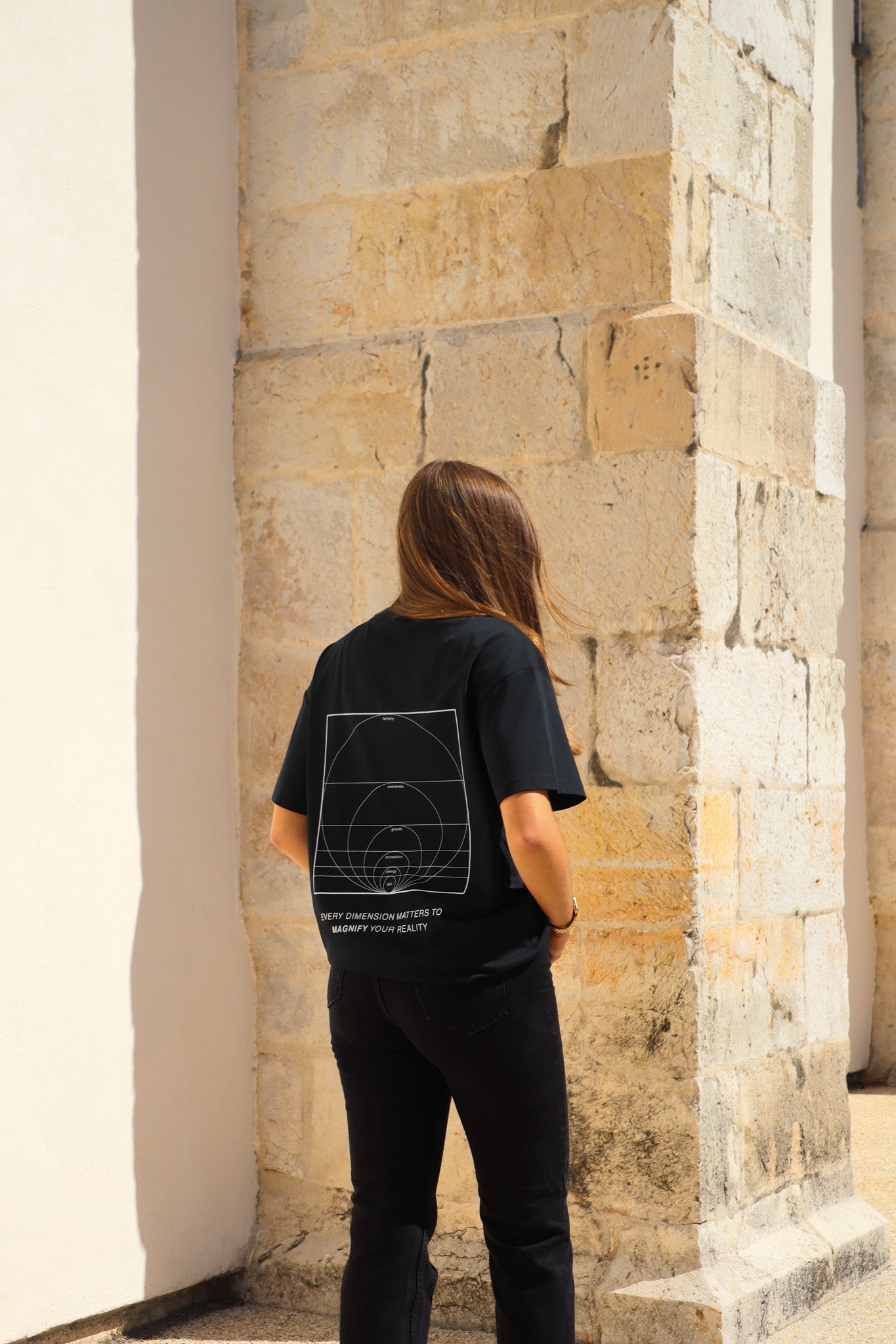 Person wearing a black t-shirt with a geometric design, standing against a stone wall.