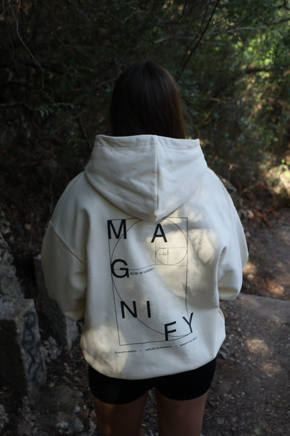 Person wearing a white hoodie with 'MAGNIFY' printed on the back in a forest setting