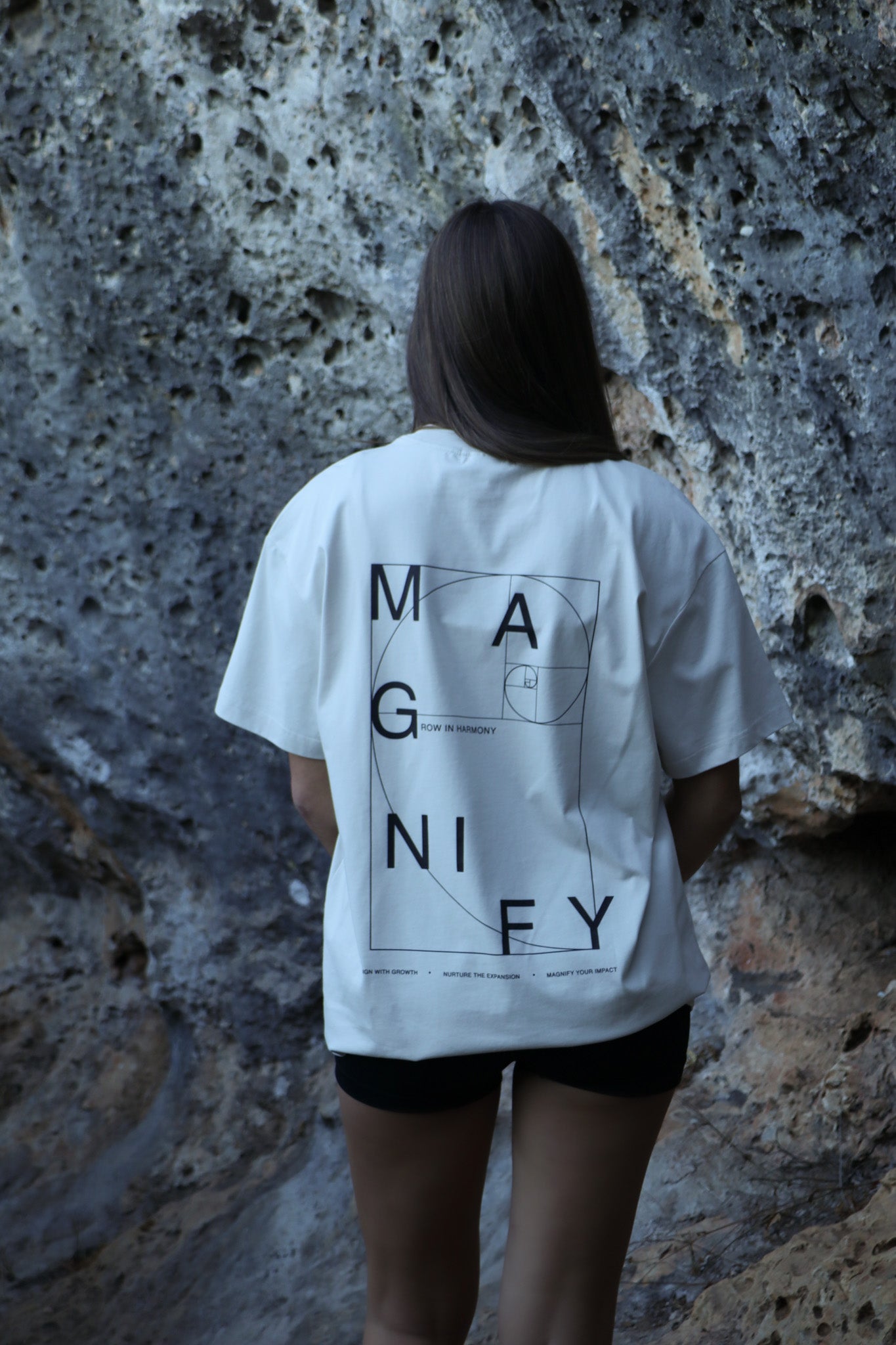 Person wearing a white t-shirt with 'MAGNIFY' design against a textured wall.