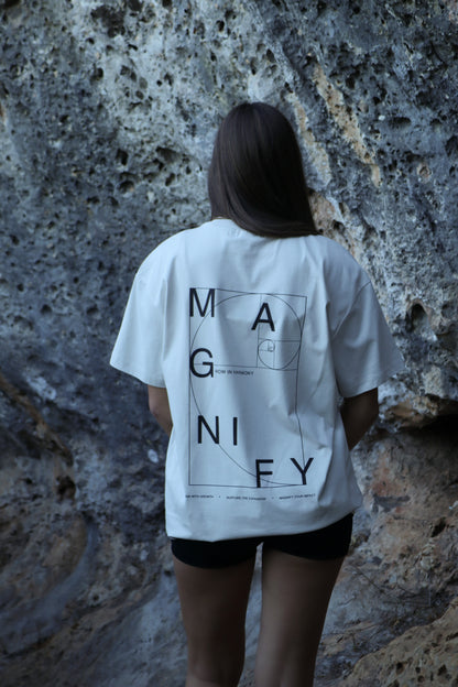 Person wearing a white t-shirt with 'MAGNIFY' design against a textured wall.