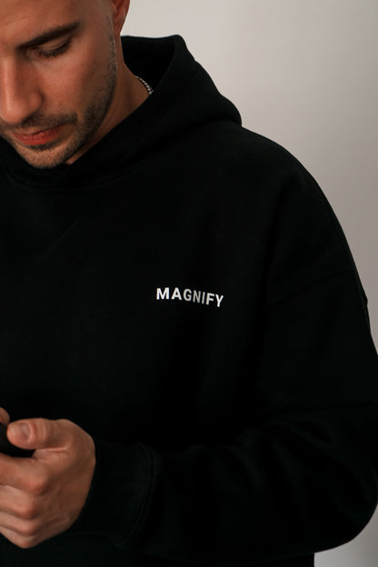 Man wearing a black hoodie with 'MAGNIFY' text on a plain background