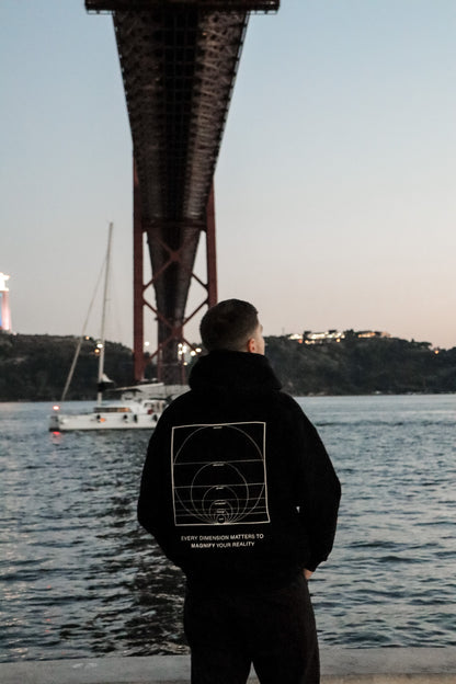 Person wearing a black jacket with a design and text, standing by a waterfront with a bridge in the background.