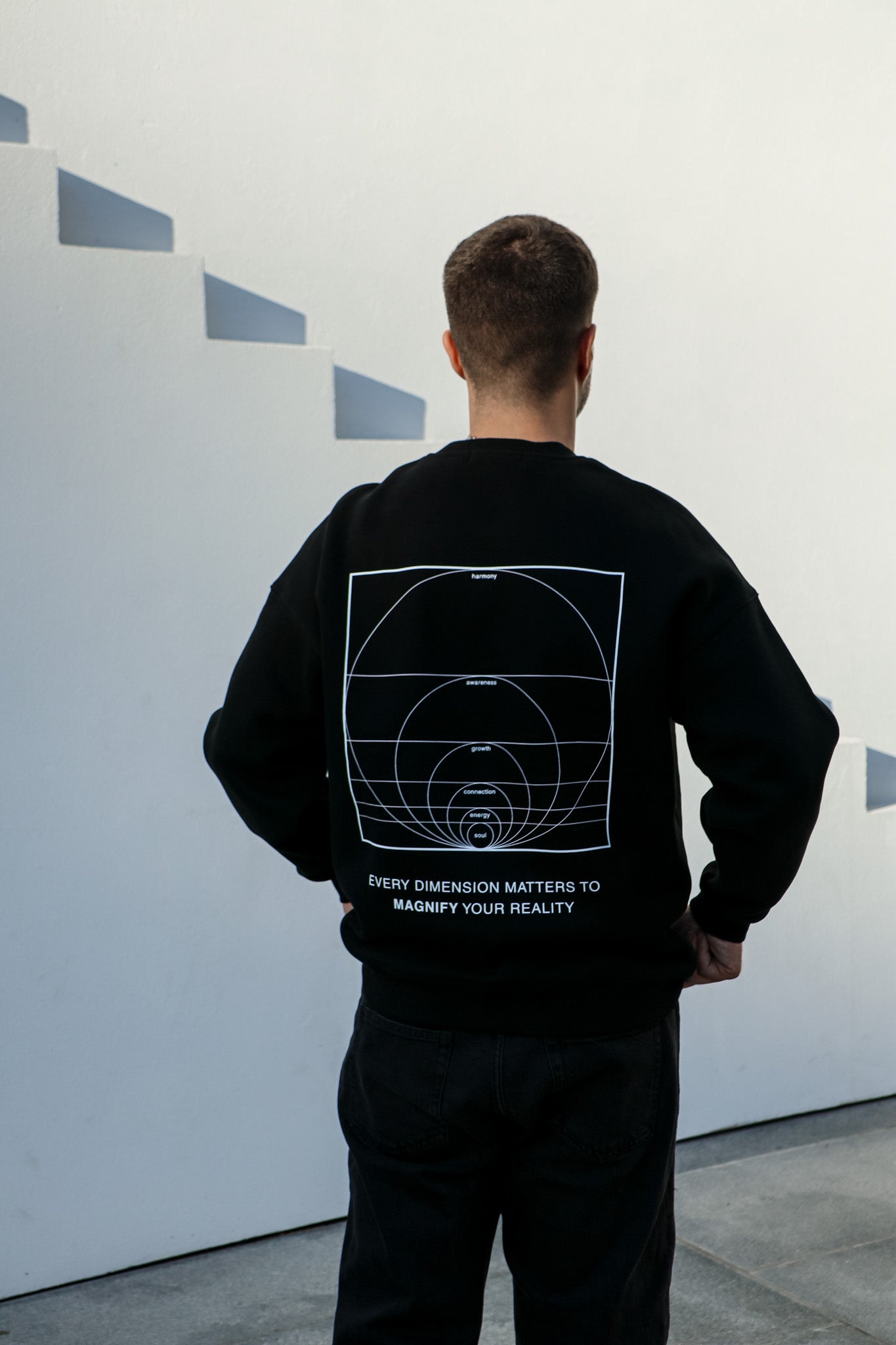 Person wearing a black sweatshirt with geometric design and text on the back, standing against a white wall.