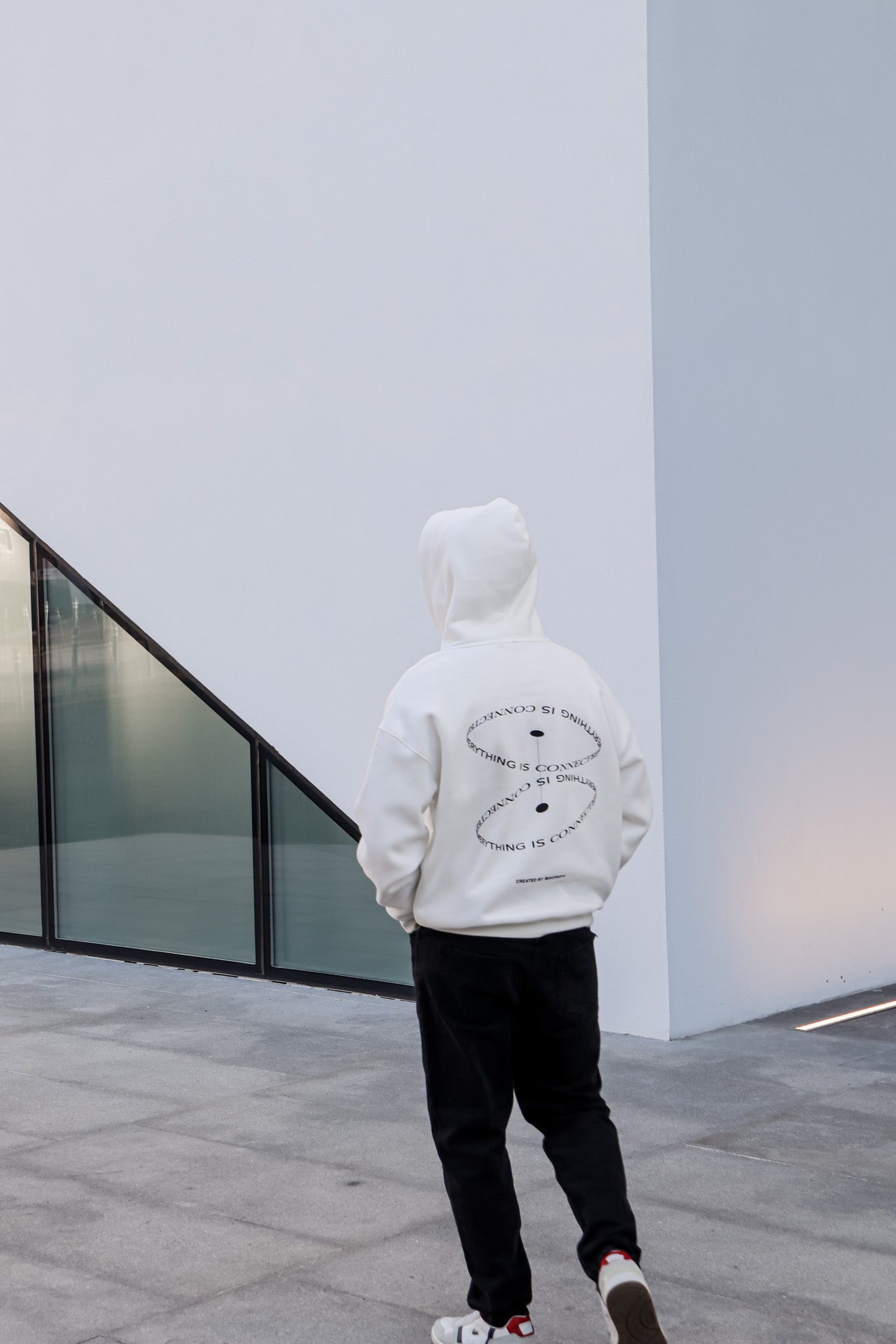 Person wearing a white hoodie with a black design, standing in front of a modern building.